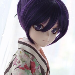 Puede incluir: Una mu&ntilde;eca con el pelo morado peinado en un mo&ntilde;o con una trenza. La mu&ntilde;eca lleva un kimono japon&eacute;s tradicional con un estampado floral y un cuello a rayas blanco y negro. Los ojos de la mu&ntilde;eca son morados y est&aacute; mirando hacia un lado.