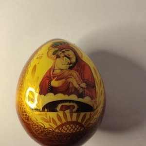 May include: A decorative egg with a religious icon of a woman and child. The egg has a yellow background with gold accents and a brown base. The icon features red, white, and gold colors.
