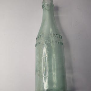 May include: A vintage, clear glass bottle with a light green tint. The bottle has a long neck and a rounded body with horizontal lines. The text "AUTO WATER" and "AMERICA'S PHYSIC" is embossed on the bottle.
