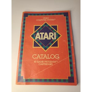 May include: An orange Atari Video Computer System catalog featuring a central blue and green diamond logo. The text reads Atari Video Computer System, Catalog, and 45 Game Program Cartridges. The cover has a dark blue and green checkered border.