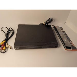 Pode incluir: Um leitor de DVD LG preto com bandeja de disco, cabo de alimentação e controlo remoto na embalagem original. O dispositivo possui uma porta HDMI, uma porta USB e uma ranhura de carregamento frontal. Cabos RCA com fichas amarela, branca e vermelha.