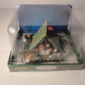 May include: A miniature camping scene inside a clear plastic display case. The scene features a green tent with a red accent, surrounded by miniature trees and figures. The background depicts a blue sky and water.