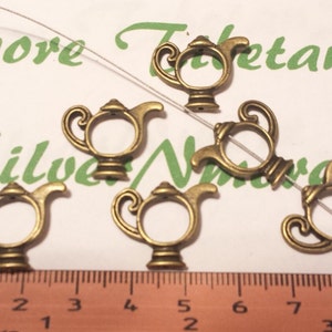 Puede incluir: Seis encantos de tetera de color dorado antiguo con un diseño decorativo. Los encantos están dispuestos sobre una superficie blanca con una regla para la escala.