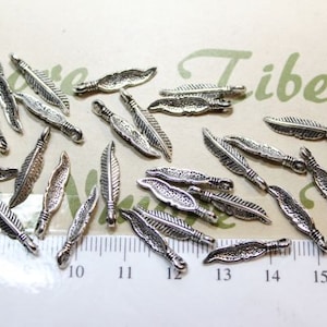 Puede incluir: Una colección de abalorios de plumas plateadas con un bucle en la parte superior para sujetar a joyas u otras manualidades. Los abalorios están dispuestos sobre una superficie blanca con una regla para la escala.