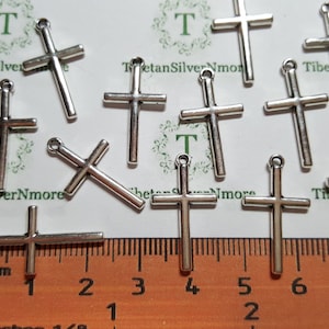 May include: A collection of silver-colored cross charms with a small loop at the top for stringing. The crosses are displayed on a white surface with a ruler showing inches and millimeters. The text "Tibetan SilverNmore" is visible.