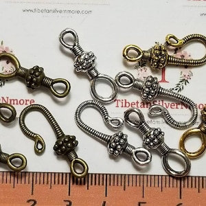 May include: A collection of decorative metal hook clasps in various finishes, including antique brass and silver. Each clasp features a decorative beaded accent and a looped end for attachment. A ruler with millimeter markings is in the foreground.