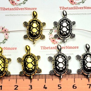 May include: Five turtle charms, two gold and three silver, are arranged on a ruler. The ruler is marked in millimetres and inches. The charms are approximately 2.5 cm in size.