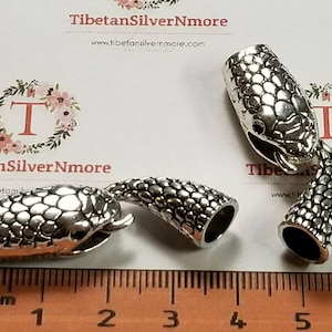 May include: Three silver snake head end caps for cords or straps. The end caps are approximately 2 cm long and feature a textured snake skin design. The end caps are displayed on a white surface with a ruler for scale.