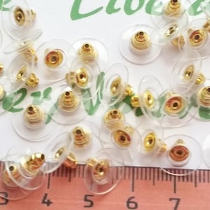 May include: A close-up of a group of 25 gold-tone earring backs with clear plastic backing. The earring backs are arranged on a white surface with a ruler in the background.