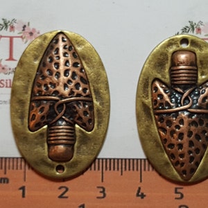 May include: Two antique-style metal pendants. Each pendant features an arrow design with a copper-coloured arrowhead and a brass-coloured oval frame. The pendants have a hole at the top for stringing.