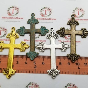 Puede incluir: Cuatro dijes de cruz de metal adornados en diferentes acabados: dorado, verde antiguo, plateado y bronce. Las cruces miden aproximadamente 5 cm de alto y presentan detalles intrincados. Los dijes se muestran en una regla para referencia de tamaño.