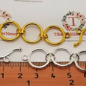 May include: Gold and silver-colored jewelry clasps with a series of connected rings. The gold clasp has a curved bar with a ball on the end. A ruler with millimeter markings is in the foreground.