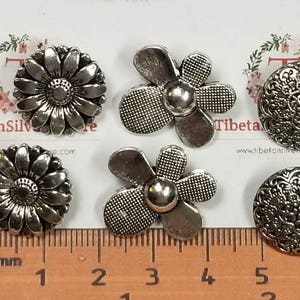 May include: Six silver-toned metal buttons with different designs.  Two buttons feature a daisy design, two buttons feature a five-petal flower design, and two buttons feature a circular design with intricate details.