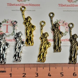 Peut inclure: Cinq pendentifs de charme en argent et en or représentant la Statue de la Liberté, chacun mesurant environ 2,5 cm de haut. Les breloques sont sur une surface blanche avec une règle pour l'échelle.