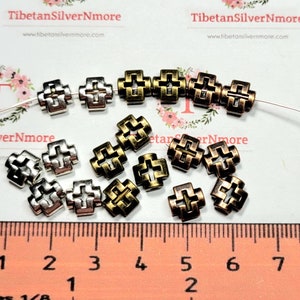Puede incluir: Una colecci&oacute;n de 16 cuentas de metal en forma de cruz, de color plateado y dorado. Las cuentas miden aproximadamente 1,2 cm de tama&ntilde;o y tienen un dise&ntilde;o calado decorativo. Las cuentas est&aacute;n dispuestas sobre una superficie blanca con una regla para la escala.