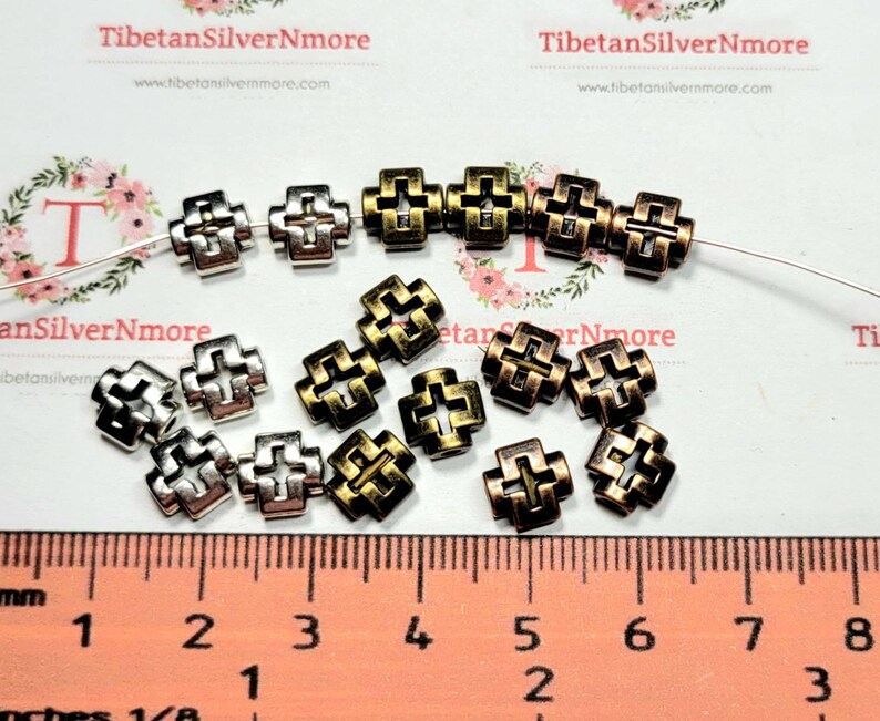 Puede incluir: Una colecci&oacute;n de 16 cuentas de metal en forma de cruz, de color plateado y dorado. Las cuentas miden aproximadamente 1,2 cm de tama&ntilde;o y tienen un dise&ntilde;o calado decorativo. Las cuentas est&aacute;n dispuestas sobre una superficie blanca con una regla para la escala.