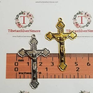 May include: Two metal crucifixes, one gold and one silver, are shown against a ruler. The crucifixes are approximately 3 inches tall.