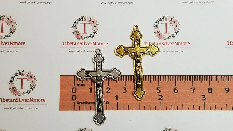 May include: Two metal crucifixes, one gold and one silver, are shown against a ruler. The crucifixes are approximately 3 inches tall.