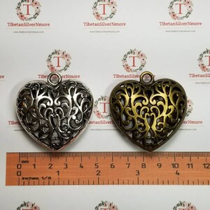 May include: Two ornate metal heart-shaped pendants, one silver and one gold, with intricate filigree designs. The pendants are approximately 2 inches in size and have a loop at the top for hanging.