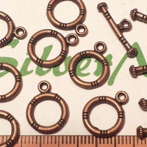 Pode incluir: Uma coleção de fechos e argolas de salto de metal com acabamento em cobre antigo. Os fechos são retangulares com um design decorativo e têm duas argolas em cada extremidade. As argolas de salto são redondas com um design decorativo e têm uma única argola numa extremidade.