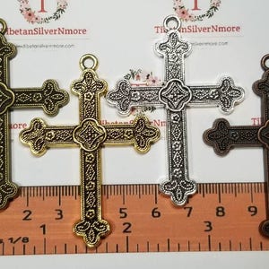 May include: Three ornate metal cross charms in gold, silver, and bronze finishes. Each cross measures approximately 2 inches in length and features intricate details. The charms are displayed against a ruler for size reference.