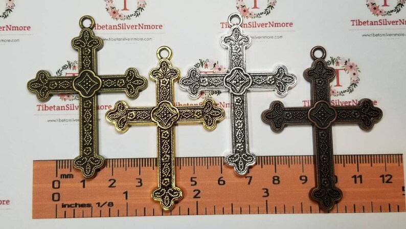 May include: Three ornate metal cross charms in gold, silver, and bronze finishes. Each cross measures approximately 2 inches in length and features intricate details. The charms are displayed against a ruler for size reference.
