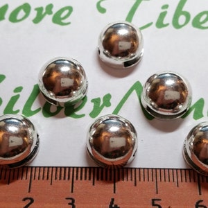 May include: Six silver metal round beads with a hole through the center. The beads are about 1 cm in diameter and are arranged in a semi-circle on a ruler.