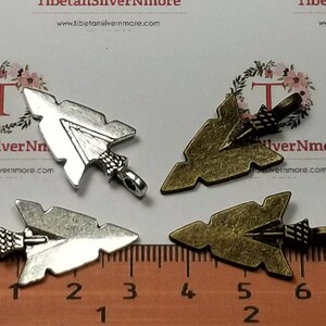 May include: Four arrowhead charms, two silver and two bronze, with a ruler for scale. The charms are about 2 inches long.