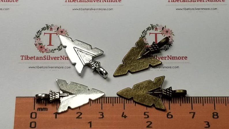 May include: Four arrowhead charms, two silver and two bronze, with a ruler for scale. The charms are about 2 inches long.
