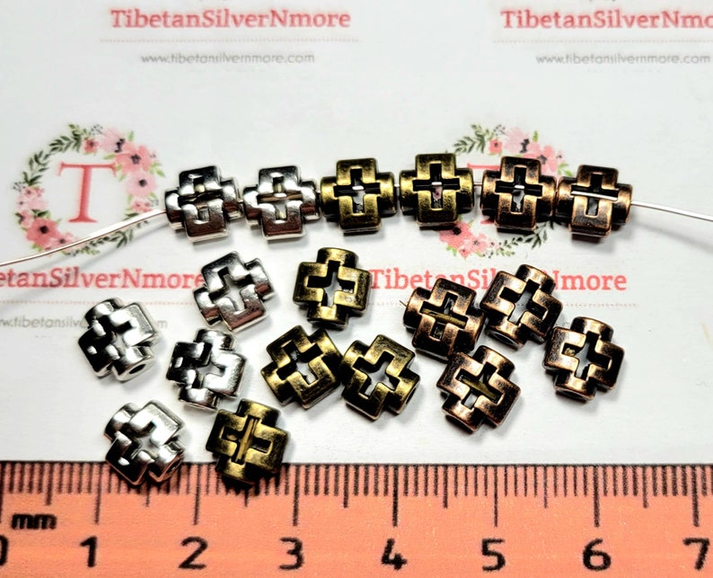 Puede incluir: Una colecci&oacute;n de cuentas de metal en forma de cruz, plateadas y bronce. Las cuentas miden aproximadamente 1 cm de tama&ntilde;o y tienen un dise&ntilde;o decorativo intrincado. Las cuentas est&aacute;n esparcidas sobre una regla con un fondo blanco.