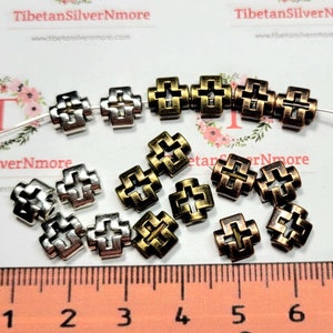 Puede incluir: Primer plano de un grupo de cuentas de metal en forma de cruz en tonos plateados y dorados. Las cuentas miden aproximadamente 1,2 cm y tienen un dise&ntilde;o decorativo intrincado. Las cuentas est&aacute;n dispuestas sobre una superficie blanca con una regla para la escala.
