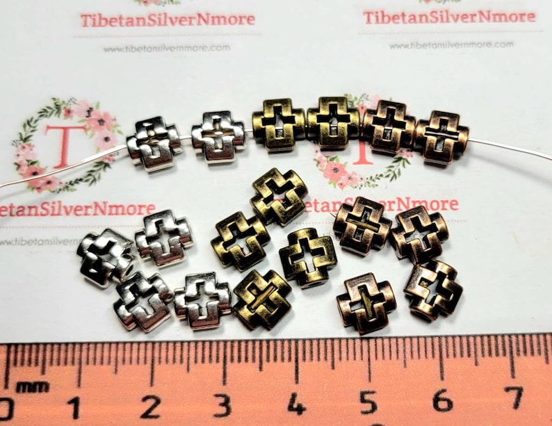 Puede incluir: Primer plano de un grupo de cuentas de metal en forma de cruz en tonos plateados y dorados. Las cuentas miden aproximadamente 1,2 cm y tienen un dise&ntilde;o decorativo intrincado. Las cuentas est&aacute;n dispuestas sobre una superficie blanca con una regla para la escala.