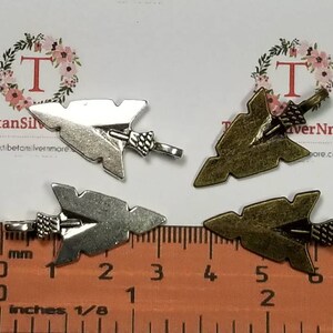 May include: Four arrowhead charms, two silver and two bronze, with a ruler for size reference. The charms are approximately 1 inch in length.