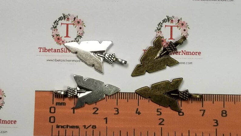 May include: Four arrowhead charms, two silver and two bronze, with a ruler for size reference. The charms are approximately 1 inch in length.