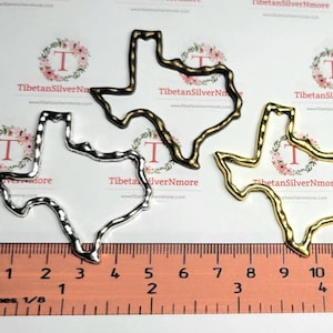 May include: Two metal Texas state outline charms, one silver and one gold, measuring approximately 3 inches in length. The charms are shown on a ruler for size reference.