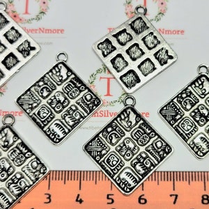 May include: Six silver-toned metal charms in the shape of a square with intricate designs. The charms have a textured surface and are approximately 2.5 cm in size.