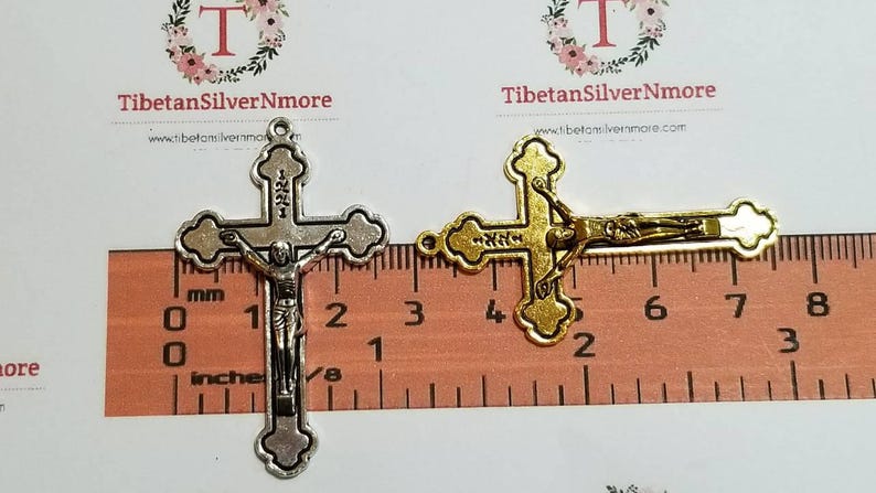 May include: Two silver and gold crucifixes with a figure of Jesus Christ on the cross. The crucifixes are measured against a ruler with inches and centimeters.