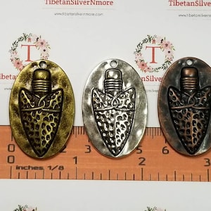 May include: Three oval-shaped metal charms with a raised arrowhead design. The charms are in antique gold, silver, and copper finishes. The charms are approximately 2.5 cm in length and are shown next to a ruler.