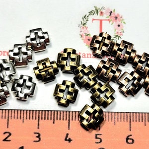 Puede incluir: Primer plano de dos juegos de cuentas de metal en forma de cruz. Un juego es plateado y el otro es dorado y cobre. Las cuentas tienen aproximadamente 1 cm de di&aacute;metro y tienen un dise&ntilde;o decorativo. Las cuentas est&aacute;n dispuestas sobre una superficie blanca con una regla en el fondo.