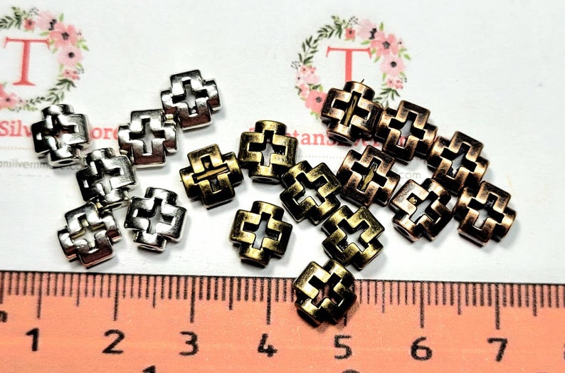 Puede incluir: Primer plano de dos juegos de cuentas de metal en forma de cruz. Un juego es plateado y el otro es dorado y cobre. Las cuentas tienen aproximadamente 1 cm de di&aacute;metro y tienen un dise&ntilde;o decorativo. Las cuentas est&aacute;n dispuestas sobre una superficie blanca con una regla en el fondo.