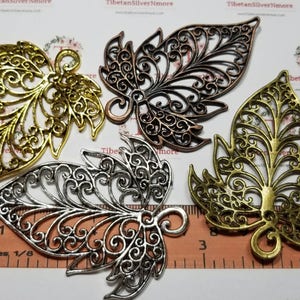 May include: Four ornate metal leaf charms in gold, silver, and bronze. The leaves are filigree and have a delicate, intricate design. The charms are approximately 3 inches in length.