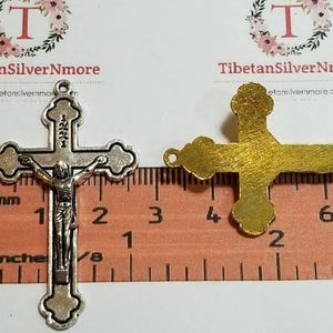 May include: Two silver-toned metal crucifixes, one with a figure of Jesus Christ, are shown next to a ruler. The ruler measures in inches and centimeters. The crucifixes are approximately 2 inches in length.