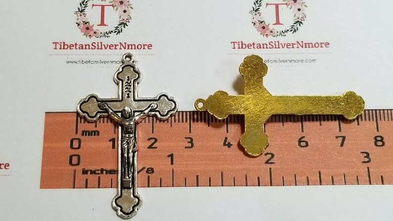 May include: Two silver-toned metal crucifixes, one with a figure of Jesus Christ, are shown next to a ruler. The ruler measures in inches and centimeters. The crucifixes are approximately 2 inches in length.