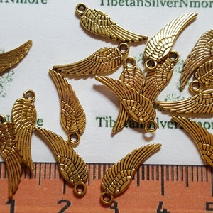 May include: A group of 15 gold-colored metal angel wing charms with a loop at the top for attaching to jewelry or other crafts. The charms are scattered on a white surface with a ruler in the bottom of the image.