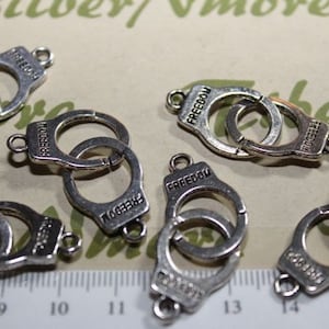 May include: Silver-toned metal charms shaped like handcuffs with the word "FREEDOM" engraved on each. The charms are arranged on a white surface with a ruler for scale.