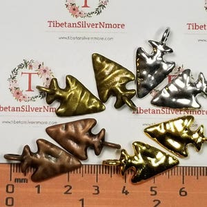 May include: Assortment of seven arrowhead-shaped metal charms in various finishes, including gold, silver, and copper. The charms are arranged on a ruler with markings in inches.
