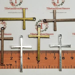 May include: Five metal cross charms in different finishes: gold, silver, bronze, and two silver. The crosses are approximately 1 inch in length and are displayed on a ruler.