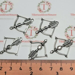 May include: Five silver-toned bow and arrow charms with a detailed design. The charms are approximately 5 cm long and have a loop at the top for attaching to jewellery or other crafts.