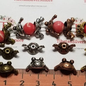 May include: A close-up of ten frog-shaped metal beads with a hole in the center. The beads are in various colors, including silver, gold, bronze, and copper. The beads are arranged on a white surface with a ruler in the foreground. The ruler is marked in inches.