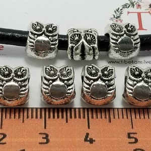 Könnte beinhalten: Sieben silberne Eulen-Perlenabstandshalter für die Schmuckherstellung. Die Perlen sind etwa 2,5 cm lang und haben ein detailliertes Eulen-Design. Die Perlen sind auf einem schwarzen Lederband und einem Lineal zur Größenreferenz abgebildet.